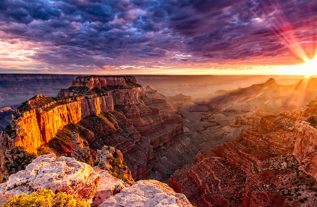 An arial photo of the Grand Canyon National Park at sunset with 'Grand Canyon Helecopter Tour' copy overlaid and dark purple clouds, bright sunlight.